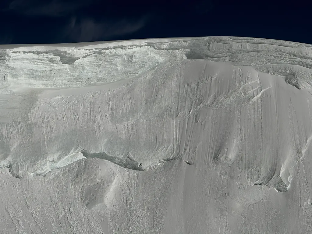 A massive, jagged ice cliff and a snow-covered mountain slope beneath a dark blue sky.