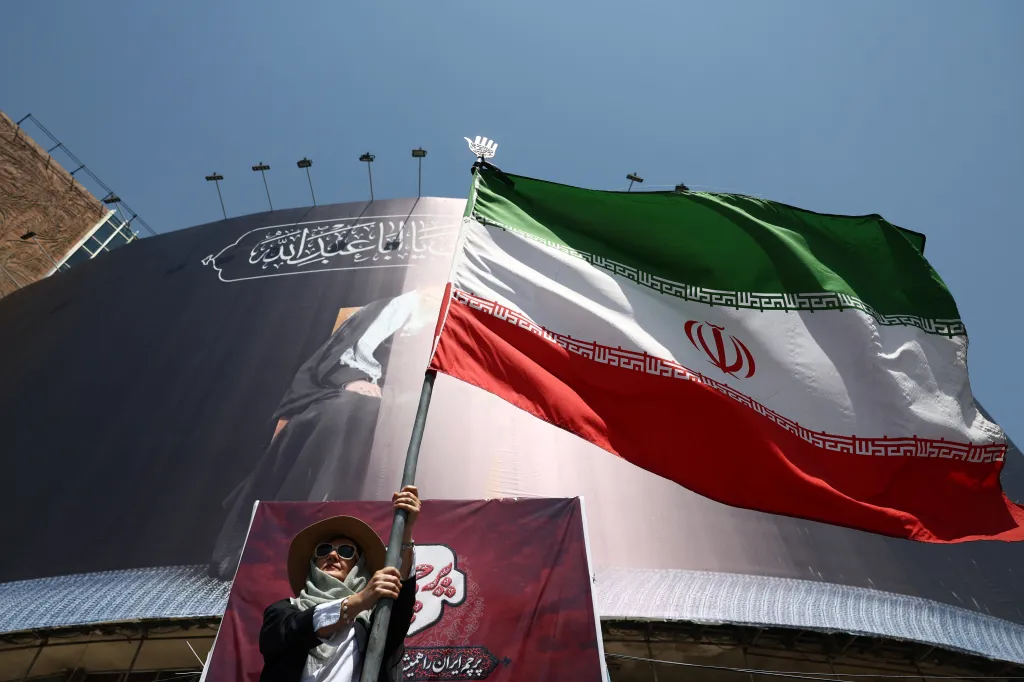 A woman holds an Iranian flag with a large banner in the background.