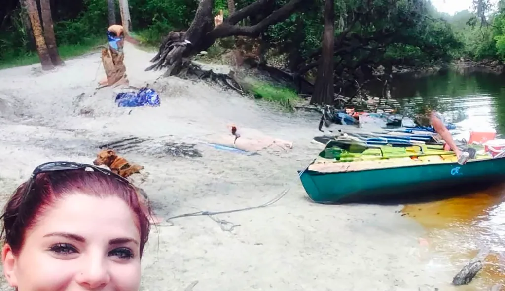 The woman is seen taking a selfie at the river with others behind her on a boat. 