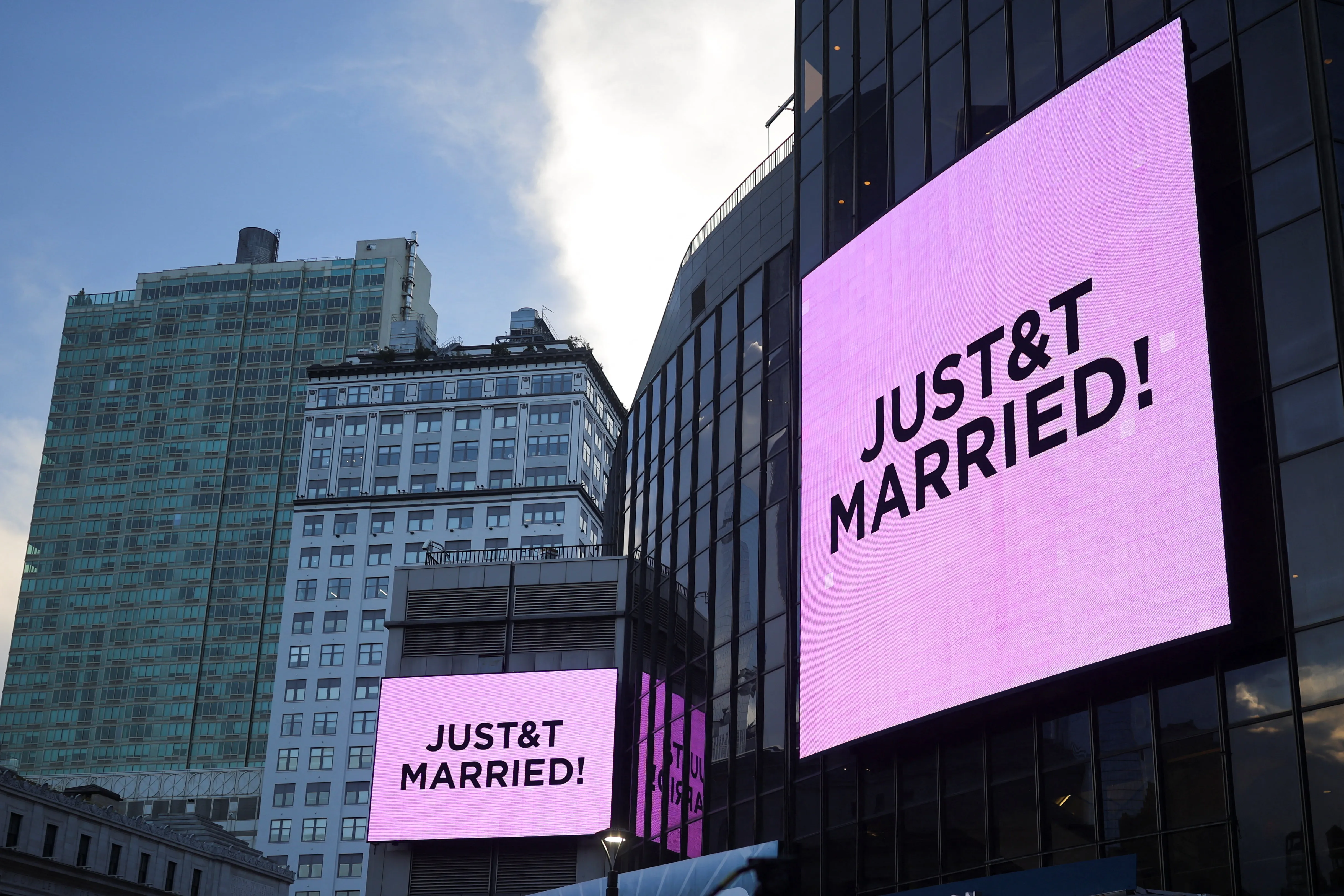 Two large digital screens on a building in New York City display "JUST & T MARRIED!" against a pink background.