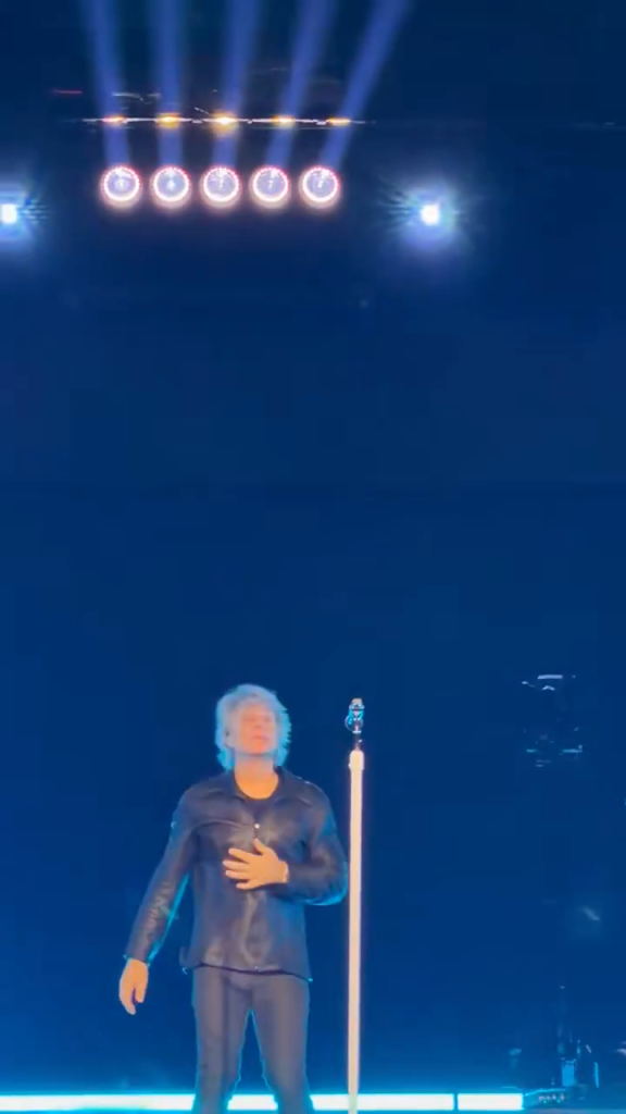 Jon Bon Jovi performs on stage with one hand on his chest, under bright blue stage lights.