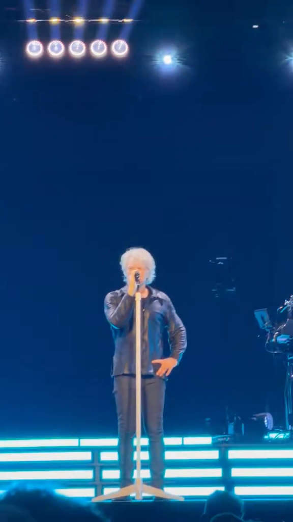 Jon Bon Jovi performing on stage, dressed in black, singing into a microphone.