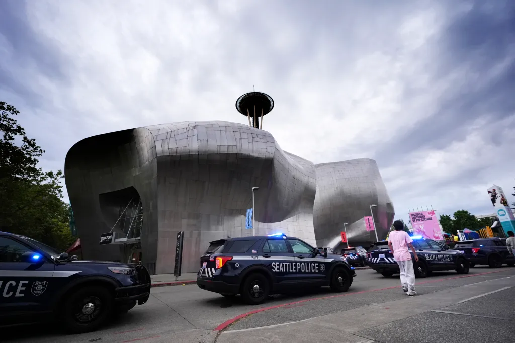 Seattle police cars with flashing lights are parked outside a modern building with the Space Needle visible in the background, responding to a shooting.