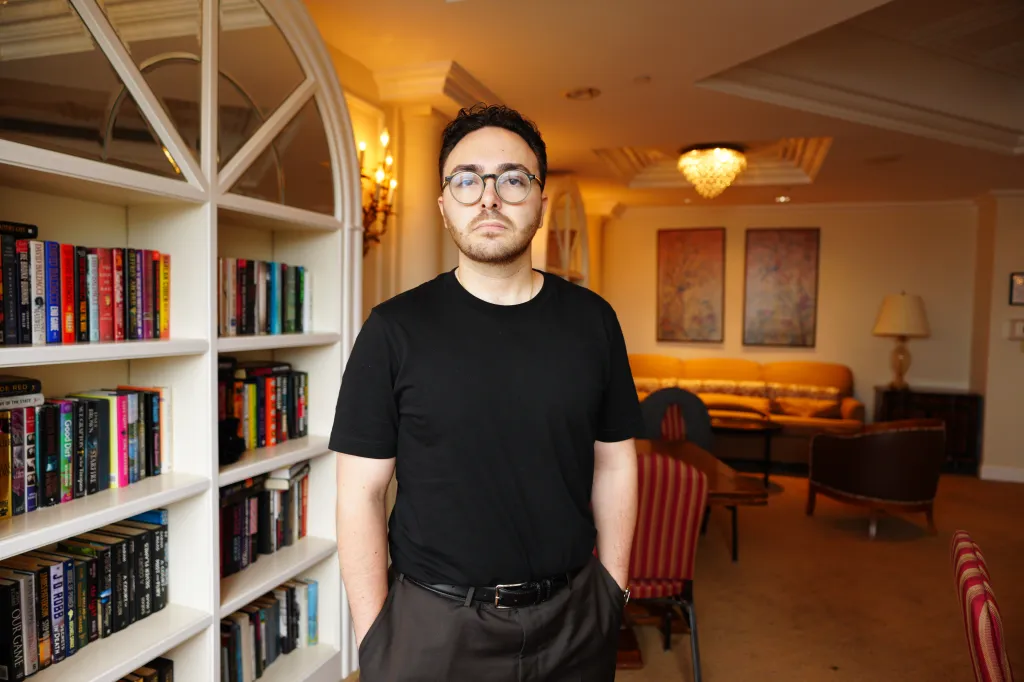 Portrait of Columbia graduate Noah Miller standing in a room with a bookshelf and living room furniture.