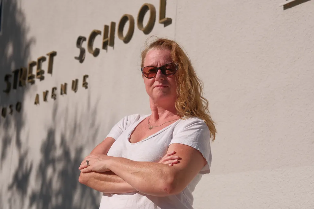 Portrait of Sonia Reiter, 48, with red hair and sunglasses, standing with arms crossed in front of a building with the words "STREET SCHOOL AVENUE" visible.