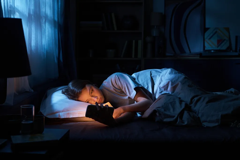 A young woman lies in bed in a dark room at night, illuminated by the glow of her smartphone.