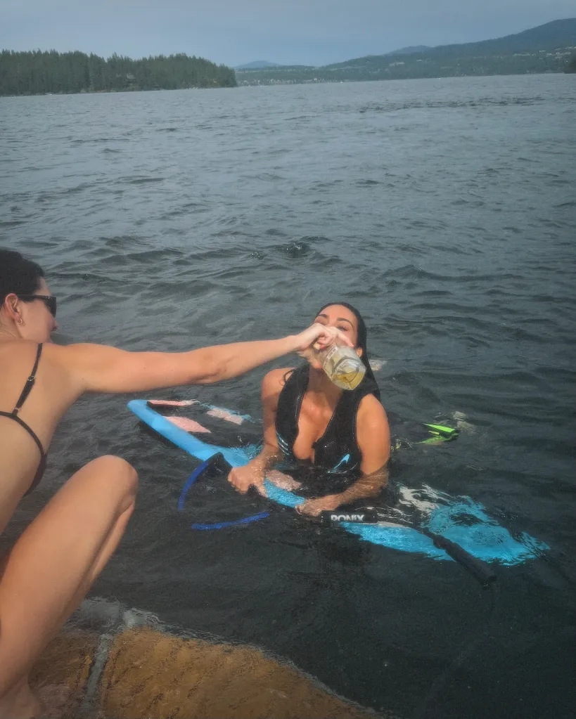 Kim Kardashian drinking from a bottle while in the water on a blue paddleboard, with a person in a black bikini reaching towards her.