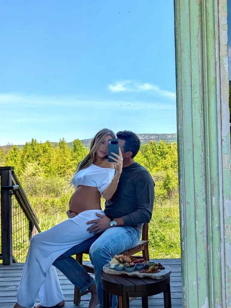 Pregnant Alex Cooper, in a white top and white pants, sitting on Matt Kaplan's lap. He's wearing a grey top and blue jeans.