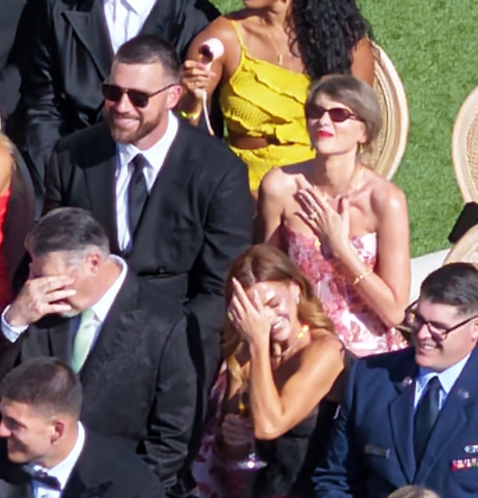 Travis Kelce smiling in sunglasses, with Taylor Swift behind him holding her hand to her chest.