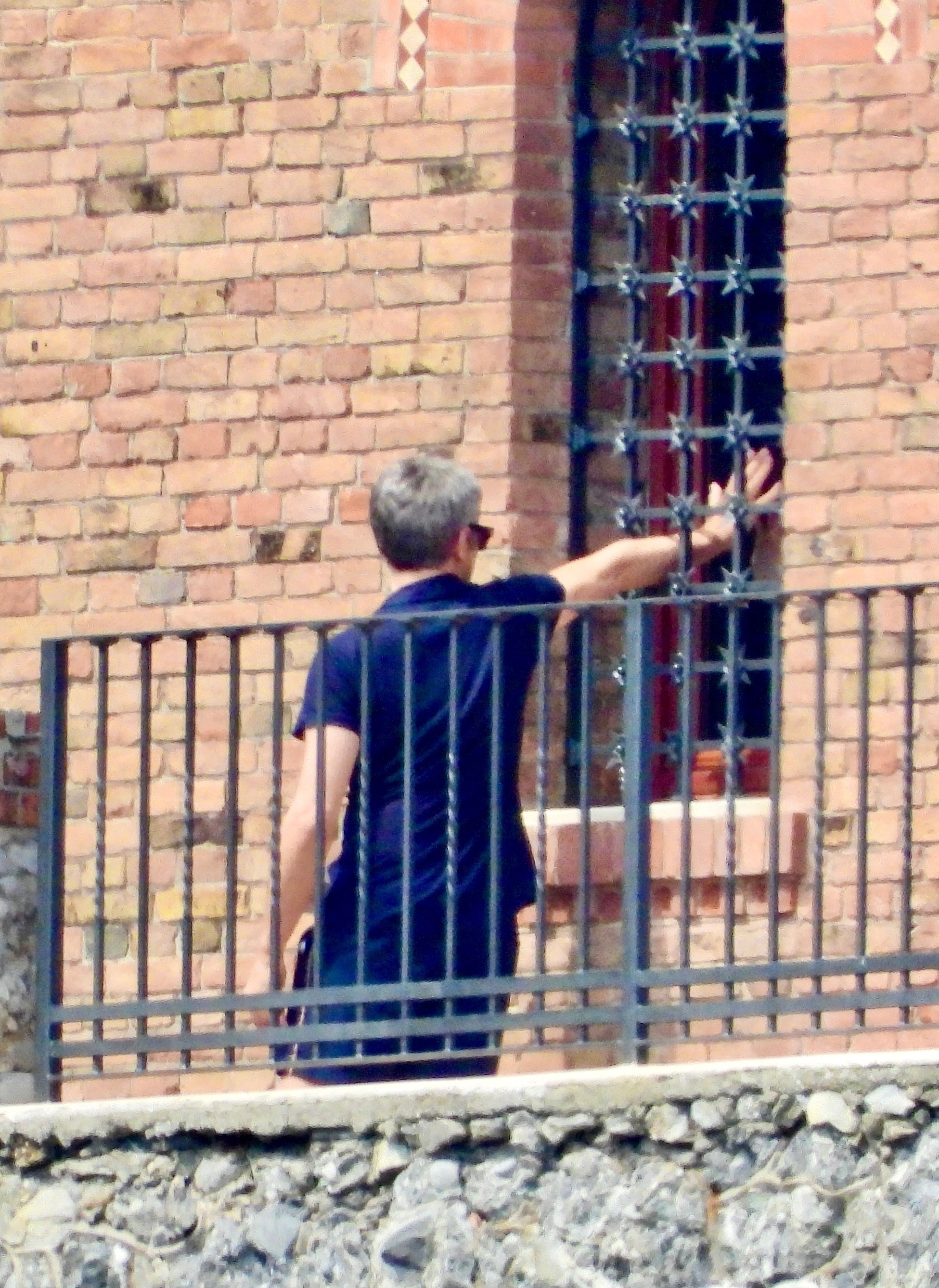 A man with gray hair in a blue shirt extends his arm through a grated window of a brick building from a balcony.