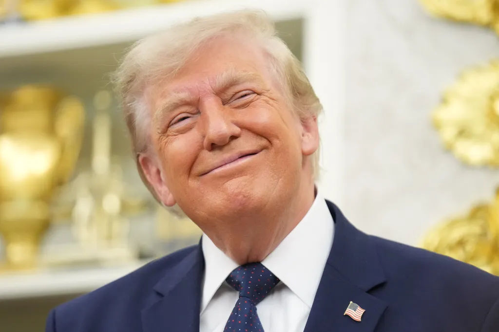 Donald Trump smiles with his head tilted, wearing a suit and tie with an American flag lapel pin.