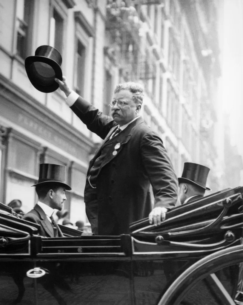 President Theodore Roosevelt waves his hat while standing in an automobile.