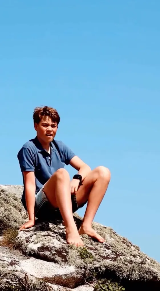 Prince George seated on a rock, wearing a blue polo shirt and shorts, looking at the camera.