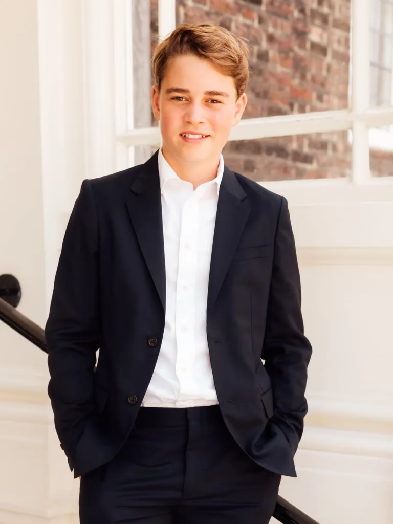 Prince George, a young boy with light brown hair, smiles at the camera, wearing a black suit and white shirt, with his hands in his pockets.