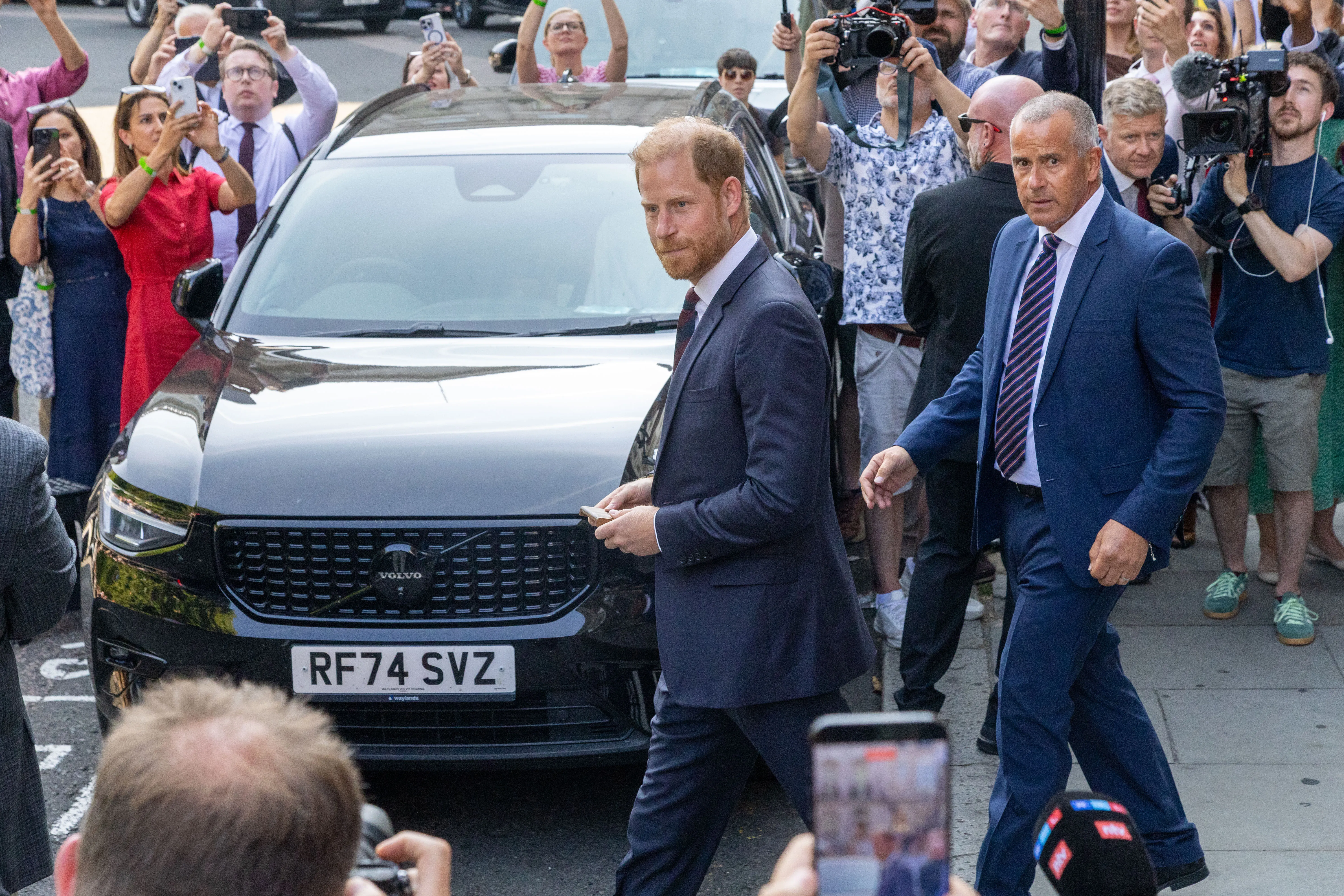 Prince Harry, Duke of Sussex, walks in front of a black car in London, surrounded by a crowd of people taking photos and videos.