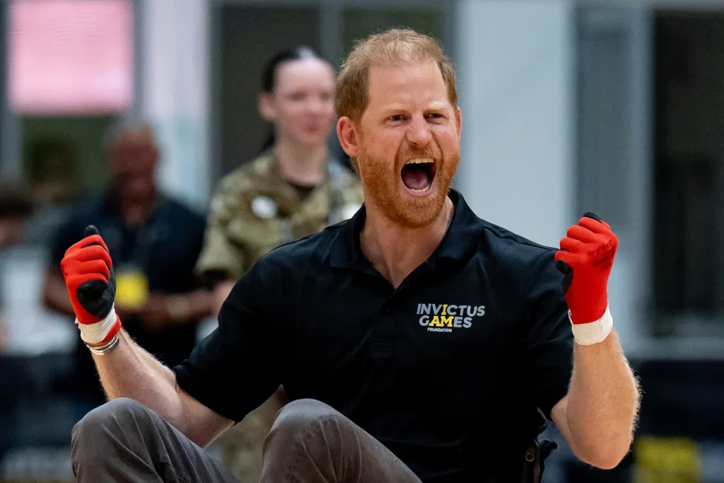 Prince Harry in a black polo shirt with "Invictus Games Foundation" logo, playing wheelchair rugby with red gloves, reacting energetically.