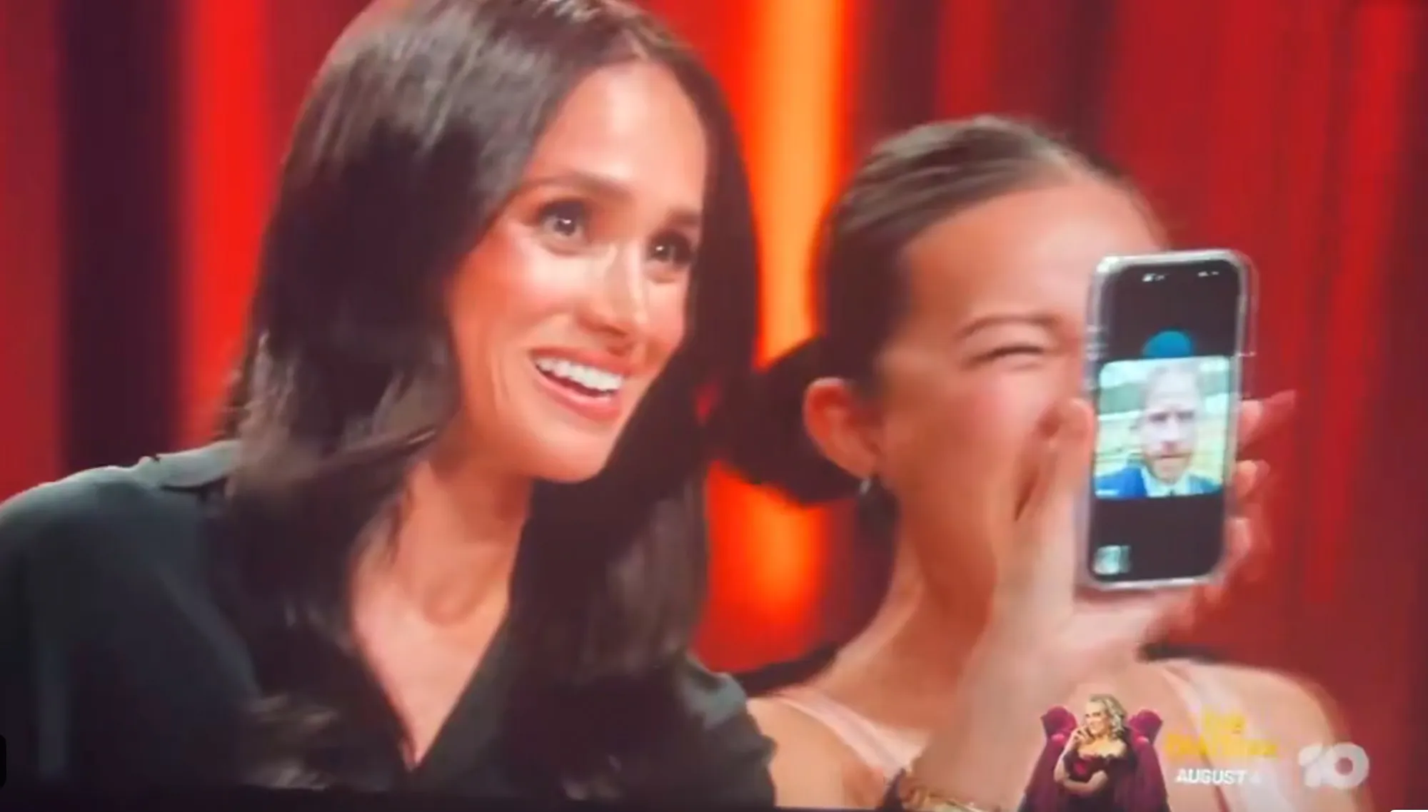 Meghan Markle smiles as a woman beside her holds up a phone with Prince Harry on video call.