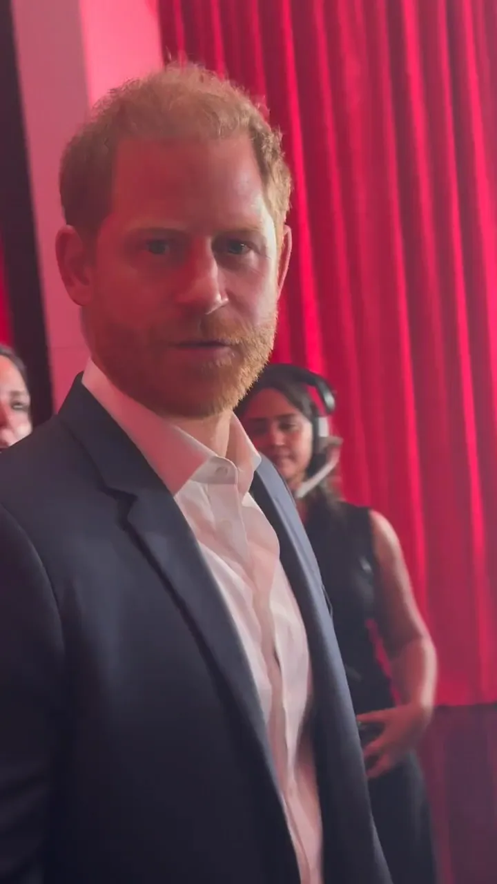 Prince Harry looking at the camera with a solemn expression, against a red curtain background.