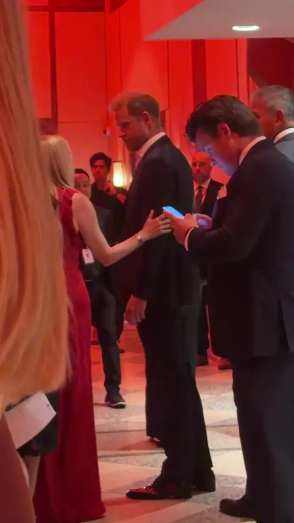 Prince Harry dressed in a black suit with a white shirt walking through a crowd of people in a red-lit room.