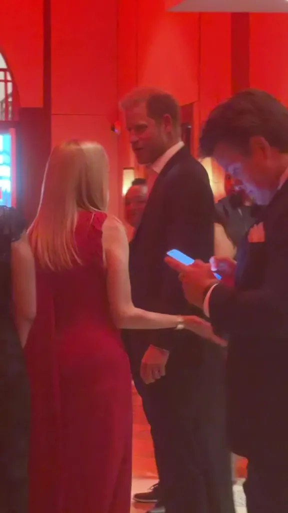 Prince Harry in a suit, with a woman in a red dress, and another man looking at his phone, in a red-lit room.