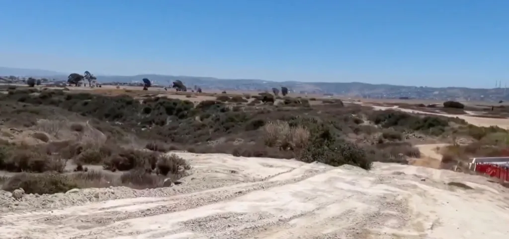 An undeveloped land parcel in Otay Mesa, California, with distant hills and scattered trees under a clear sky.