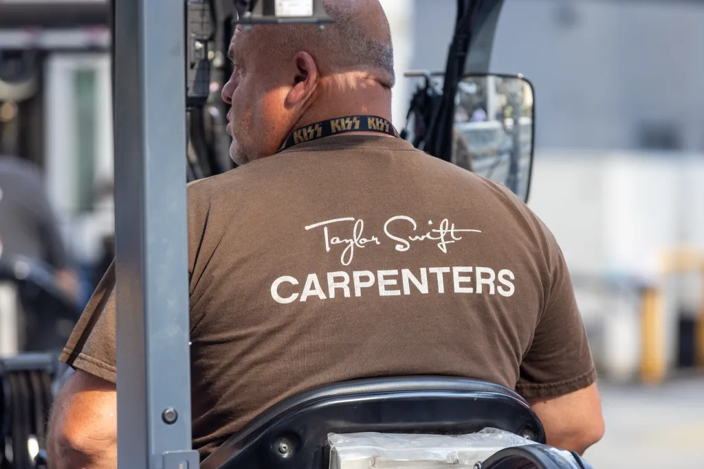 A man in a brown "Taylor Swift Carpenters" t-shirt is seen from the back, driving a forklift.