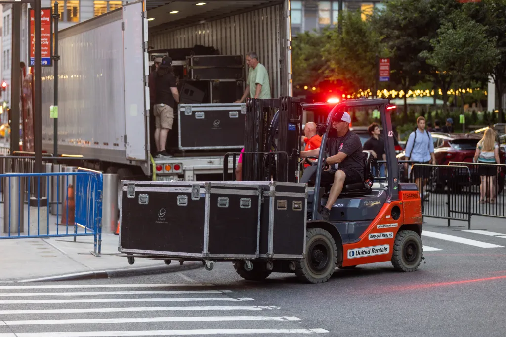 A forklift transporting props with a large truck in the background from which more props are being unloaded.