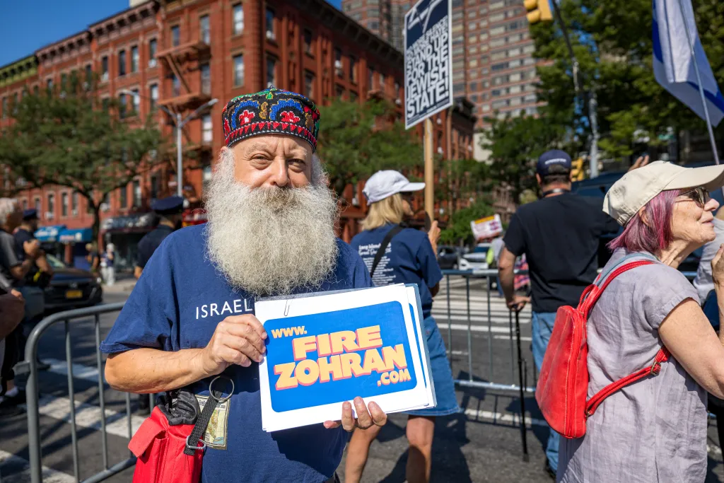 A man wearing a multi-colored embroidered cap and a full white beard holds a "www.firezohran.com" sign at a rally.
