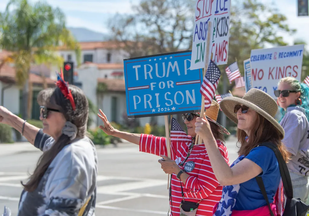 Anti-Trump protestors holding signs that read "Trump for Prison," "No Kings," and "Stop ICE Abuse & Kids in Camps."