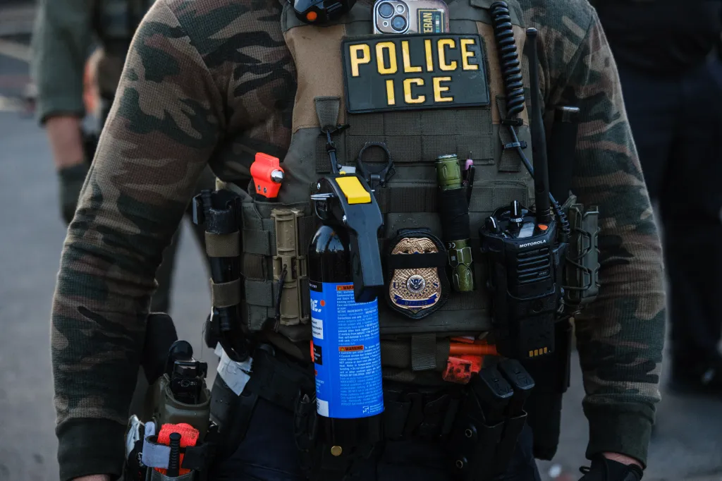An ICE officer in camouflage shirt and tactical vest, with “POLICE ICE” patch, pepper spray, radio, and badge visible.