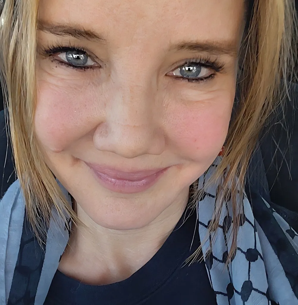 Laura Pinho, a blonde woman with blue eyes, wearing a black shirt and a patterned gray and black scarf.