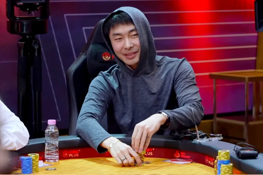 Poker player Chau Shing Lam, also known as Andrew Lam, in a grey hooded sweatshirt, smiles as he stacks poker chips at a table.