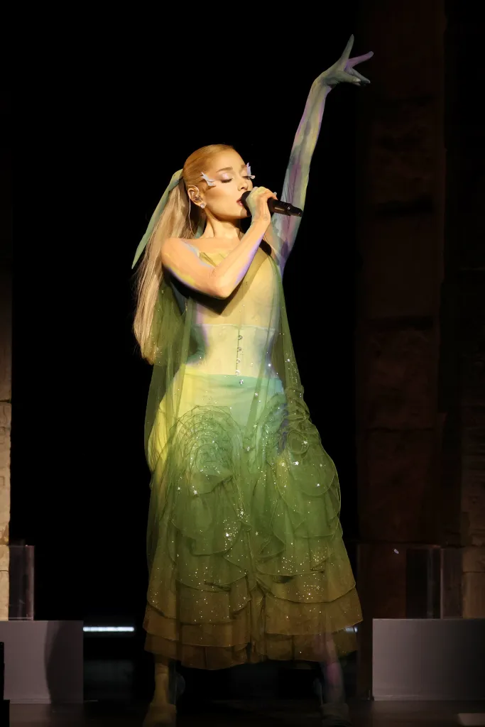 Ariana Grande performing onstage in a shimmering green gown with sheer sleeves.
