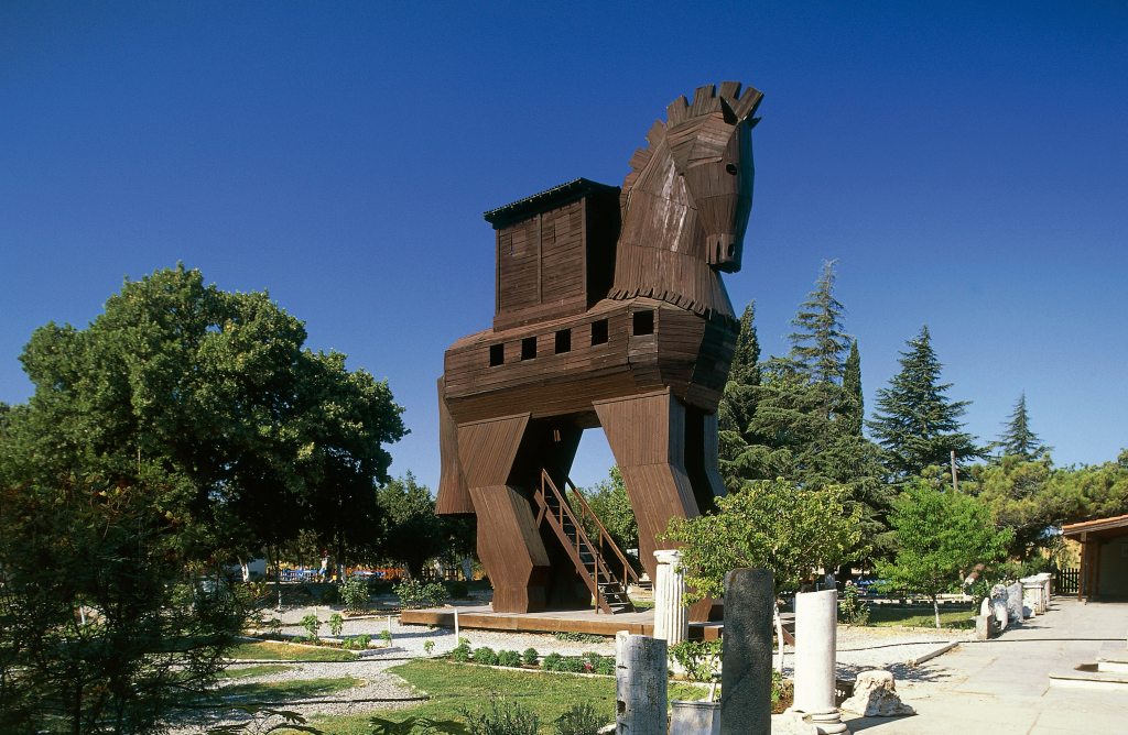 Reconstruction of the Trojan Horse in Hisarlik, Turkey, with a clear blue sky, green trees, and historical columns.