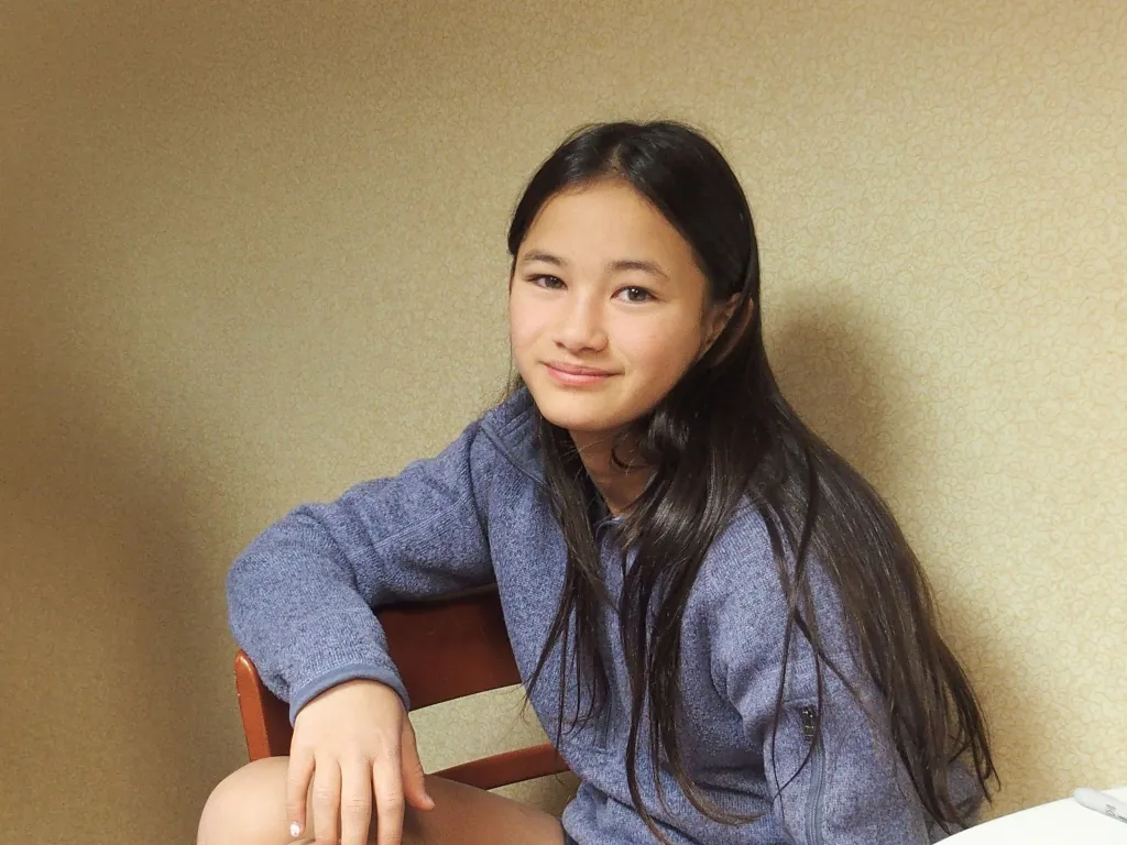 Kaylee Hottle with long dark hair, wearing a grey zipped jacket, sitting on a wooden chair and smiling at the camera.