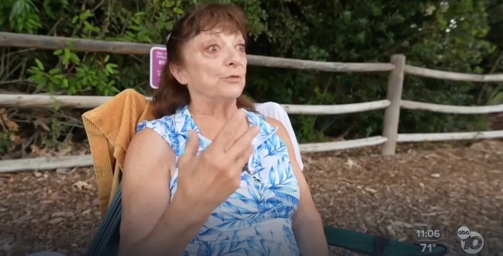 A woman with brown hair, wearing a white and blue floral top, sits in a chair and gestures with her right hand while speaking.