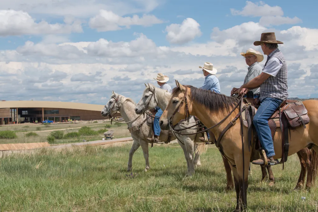 Four riders on horseback in a grassy field with the Teddy Roosevelt Presidential Library in the background.