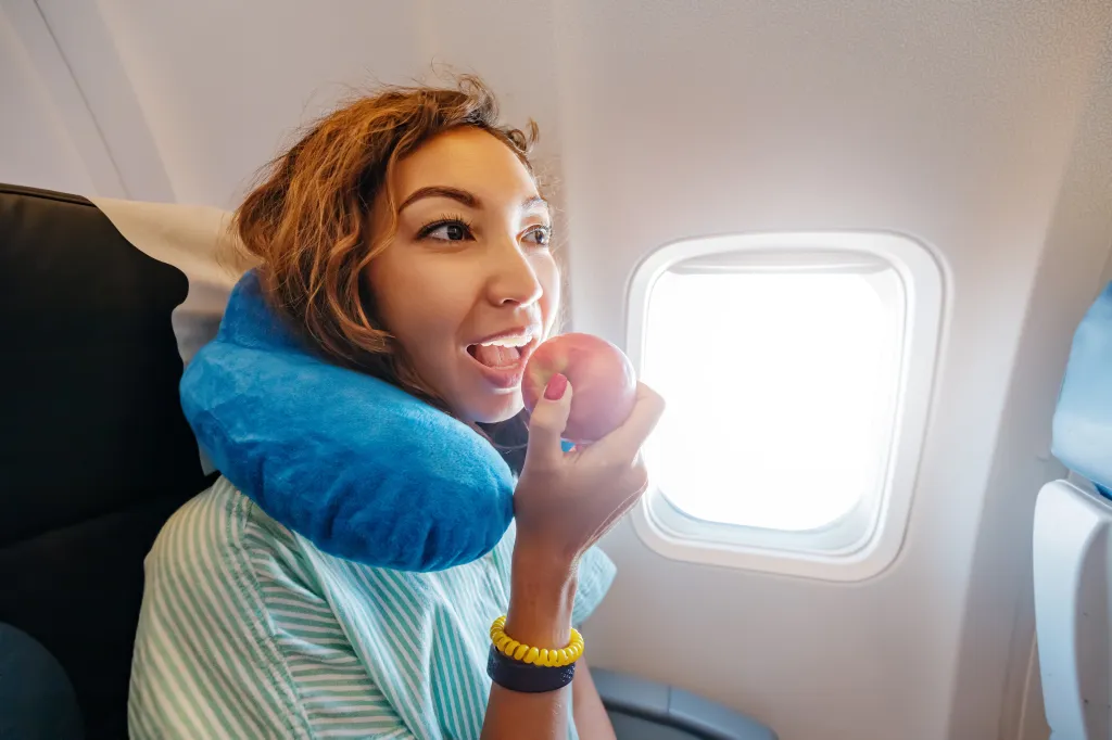 A woman wearing a neck pillow and striped shirt eating an apple in an airplane cabin.