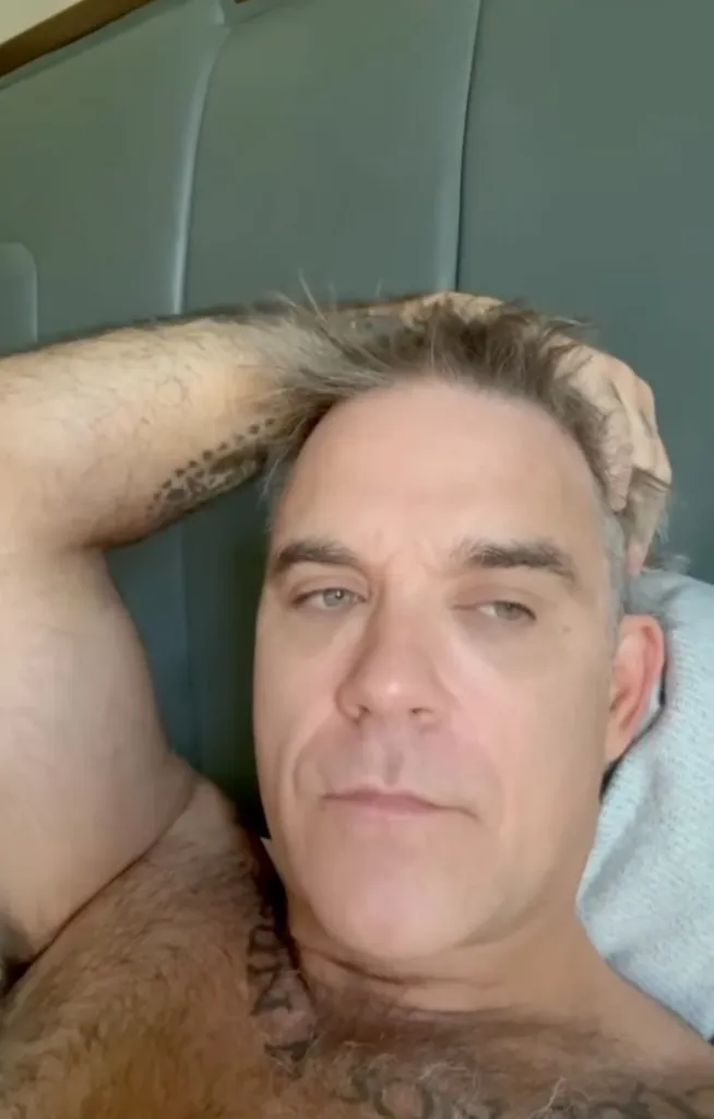 Robbie Williams lying on a pillow, shirtless, with a tattoo visible on his left arm and shoulder.