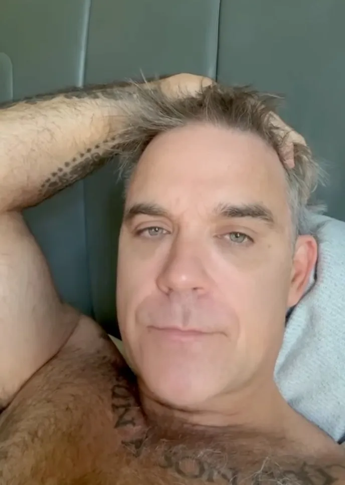 Robbie Williams wearing a messy gray hair and showing off a tattoo on his shoulder, responding to talk about a white substance on his mic.
