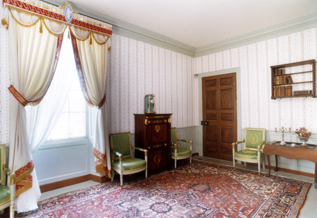 Interior of the Shirley-Eustis House with decorated curtains, patterned wallpaper, a red and blue area rug, and antique furniture.