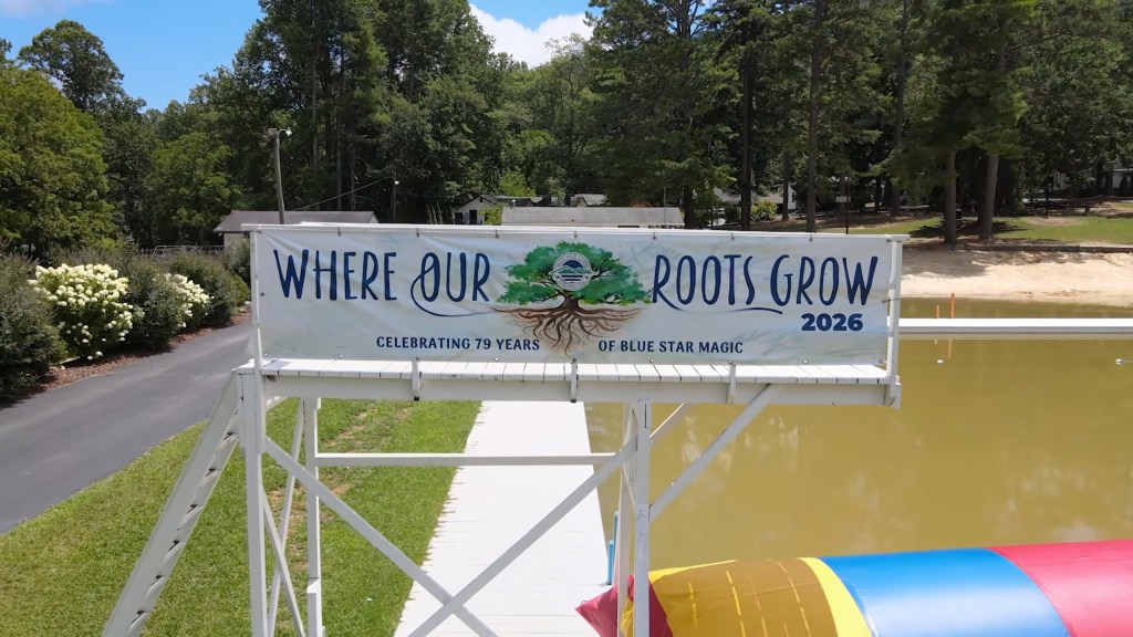A banner reading "Where Our Roots Grow 2026, Celebrating 79 Years of Blue Star Magic" hangs over a lake.