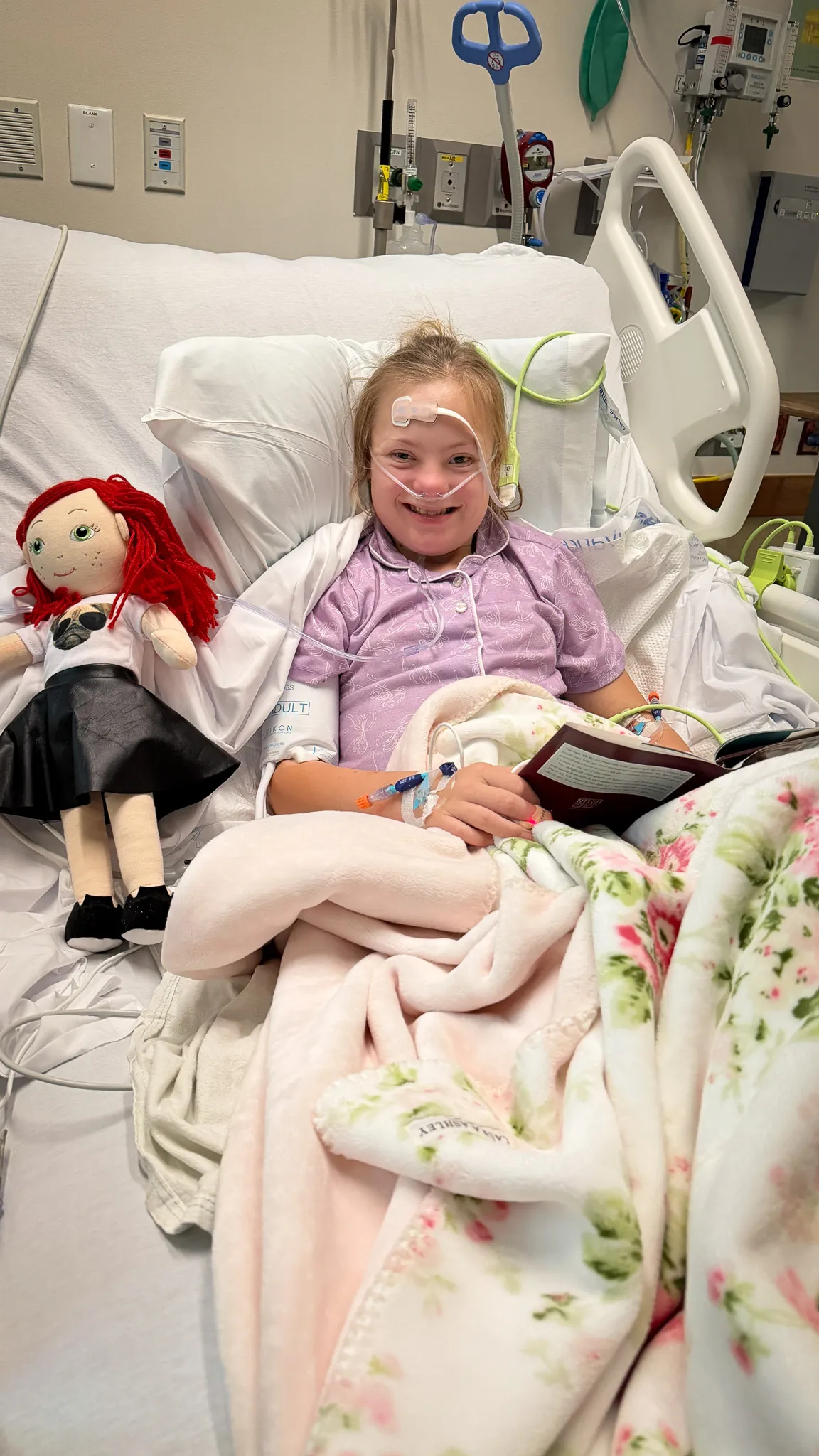 A smiling young girl in a hospital bed with a breathing tube, holding a book, with a doll beside her.