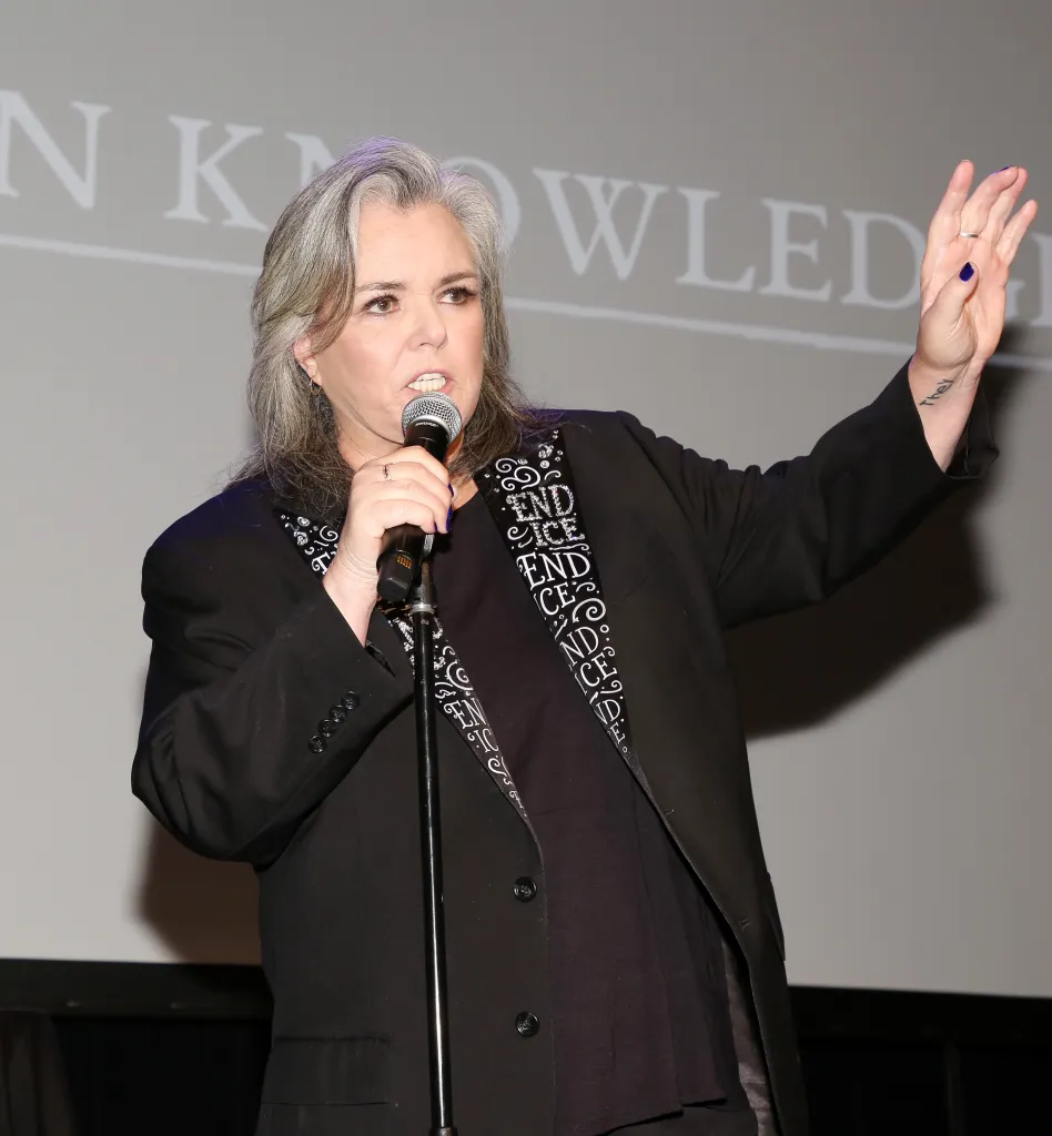 Rosie O'Donnell speaking into a microphone, with her hand raised, during