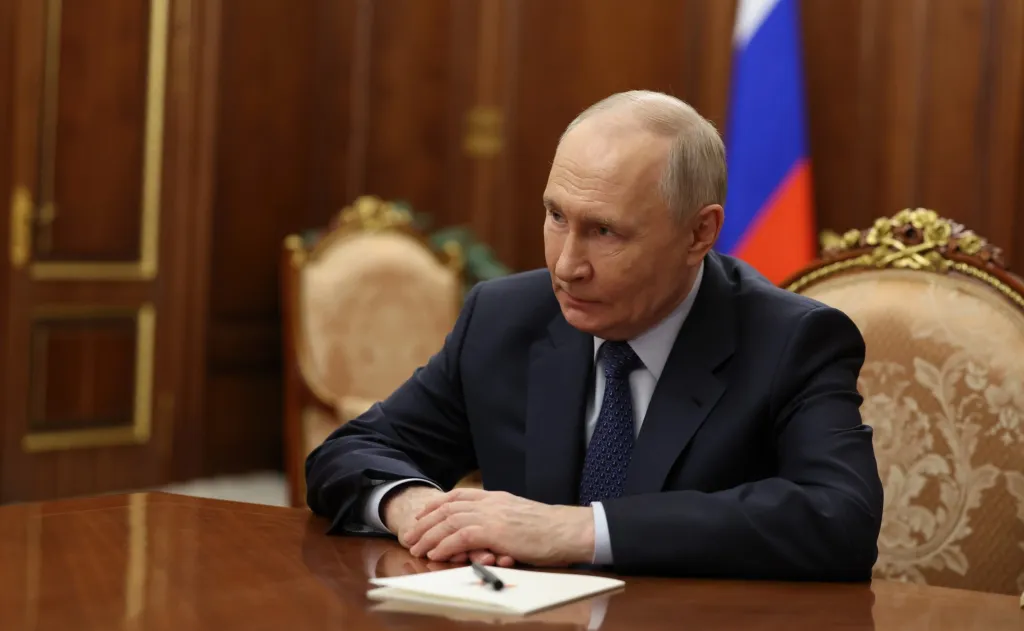 Russian President Vladimir Putin in a dark suit with his hands clasped on a wooden table, listening intently.
