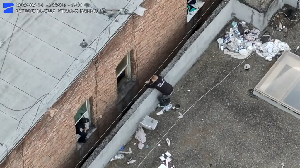 A man climbing over a narrow rooftop ledge in an alleyway during a police chase.