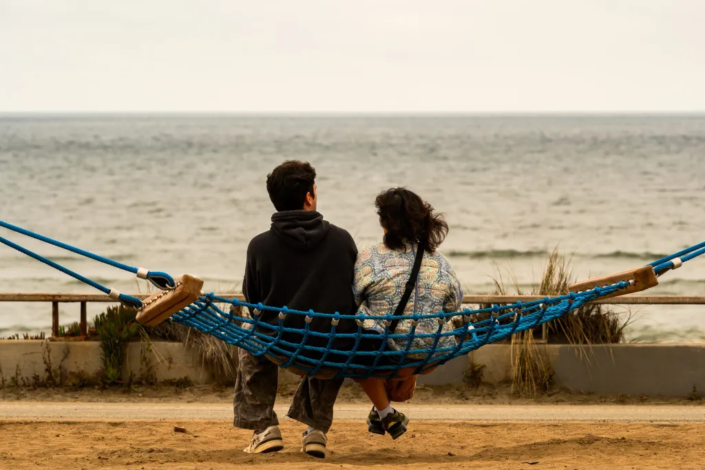Two people sitting in a blue net hammock, overlooking the ocean.