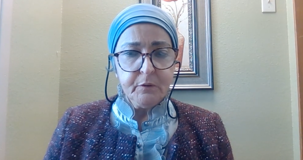 Sarwa Aldoori, a woman wearing a light blue head covering, glasses, and a brown speckled jacket over a ruffled blue shirt.