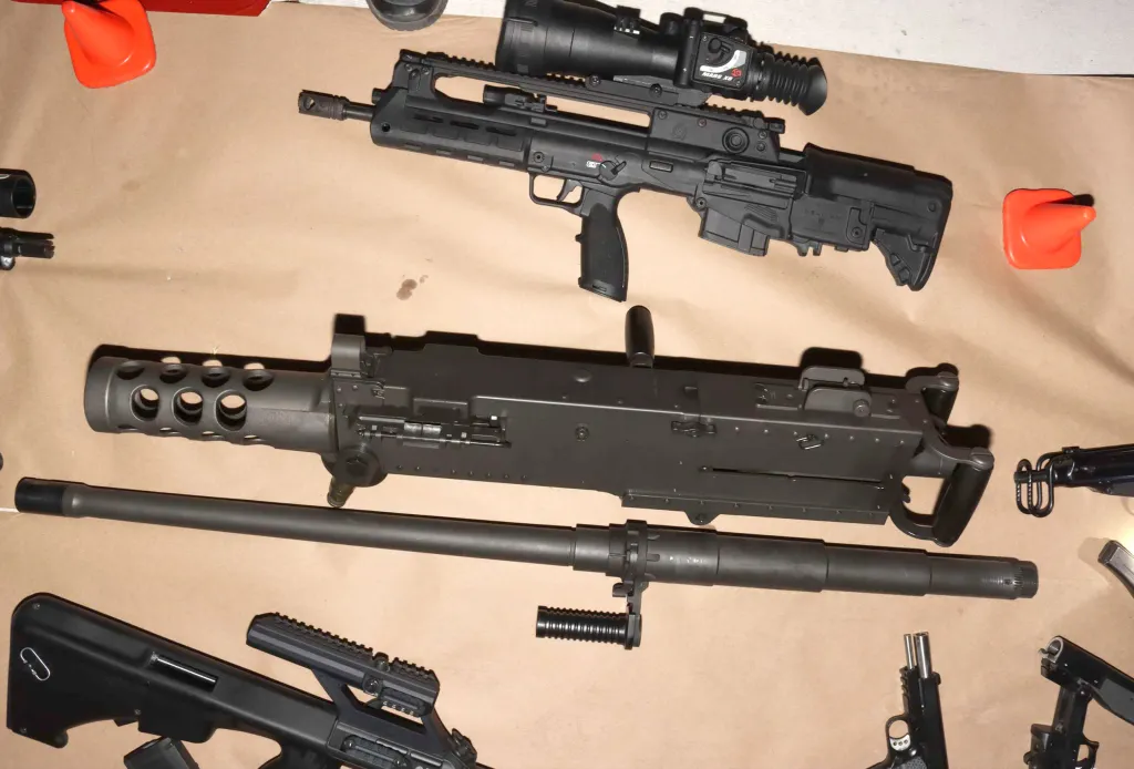 Assortment of firearms and gun parts on a light brown surface, including an assault rifle with a scope, a heavy machine gun with a barrel next to it, and handguns.