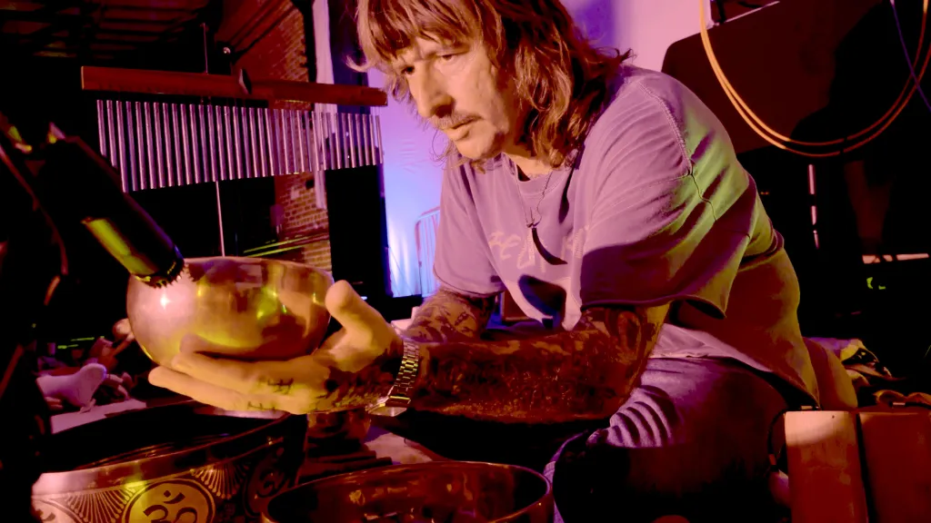 A person with long hair and a mustache holds a Tibetan singing bowl at an ambient music event.
